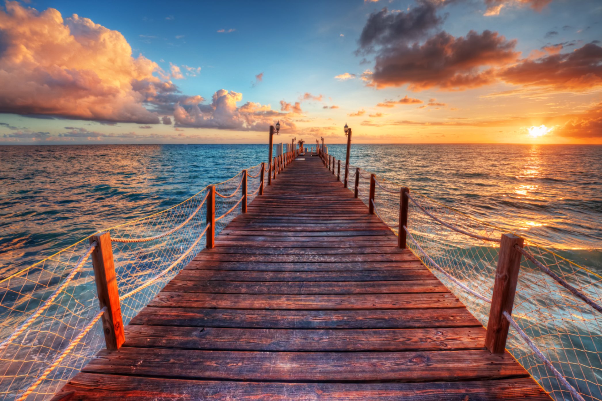 Brownish-red wooden bridge with ropes connecting its posts extends over a blue ocean at sunset. This photo represents Angela V. Fox Coaching’s expertise in moving clients from where they are to where they want to be.